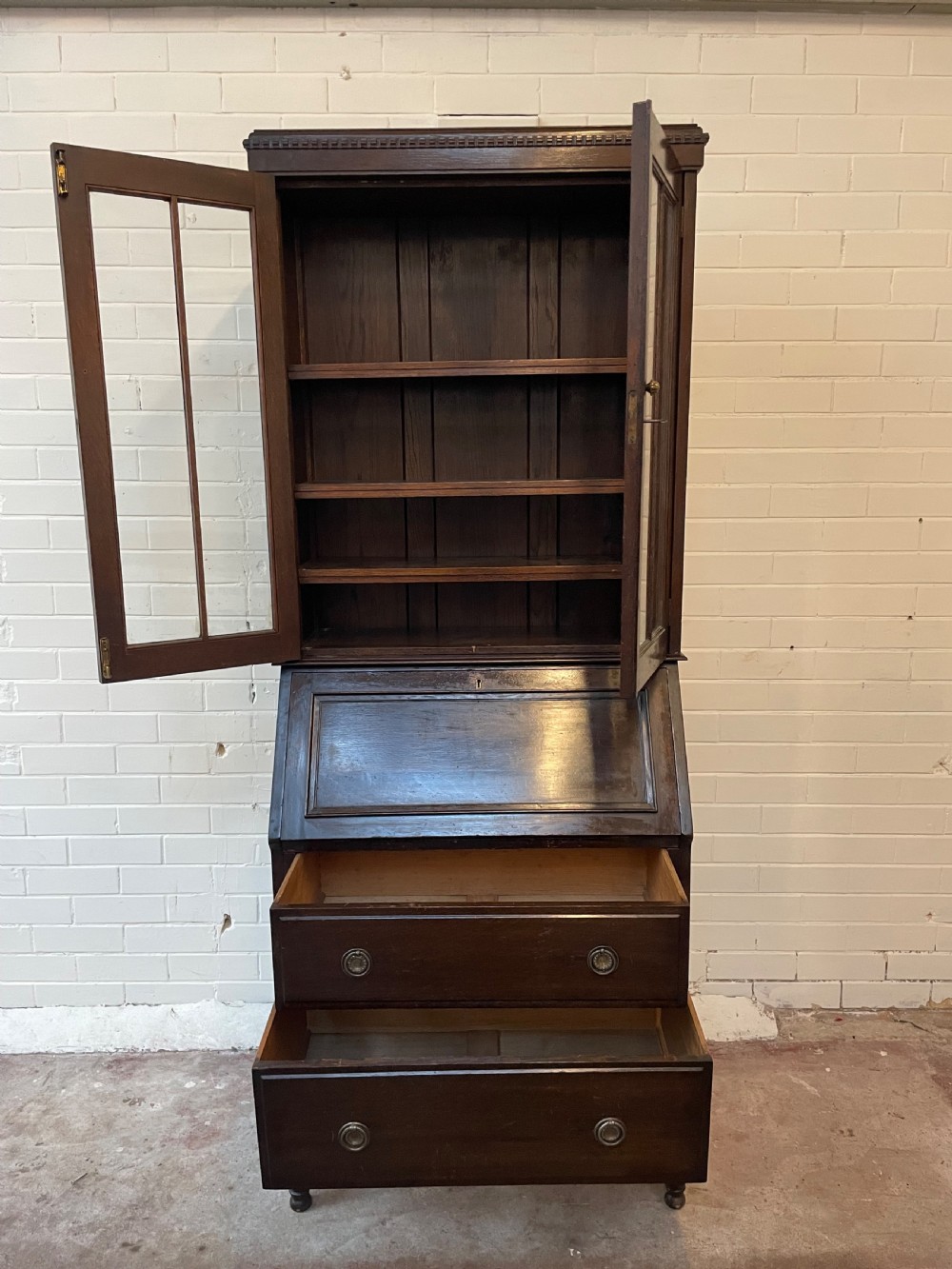 A dark oak Edwardian bureau bookcase featuring a twin-glazed door upper cabinet positioned above a slant-front writing slope and two lower drawers on turned legs. View IV
