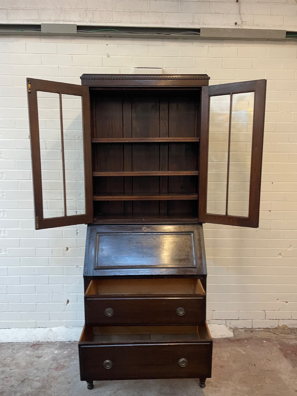 A dark oak Edwardian bureau bookcase featuring a twin-glazed door upper cabinet positioned above a slant-front writing slope and two lower drawers on turned legs. View VI