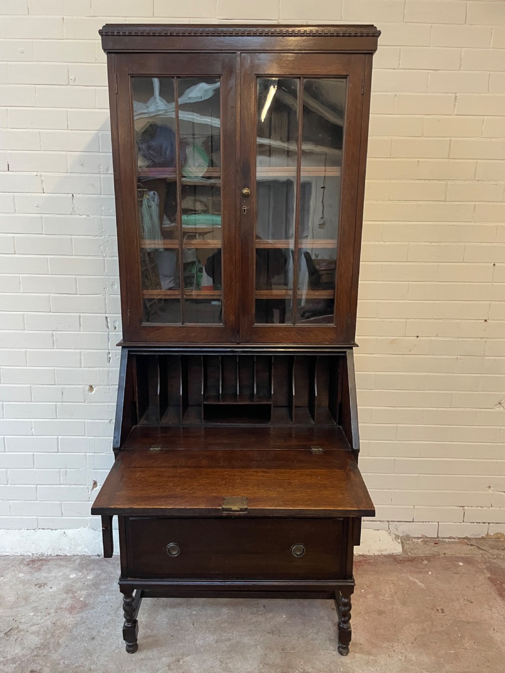 A dark oak Edwardian bureau bookcase featuring a twin-glazed door upper cabinet positioned above a slant-front writing slope and two lower drawers on turned legs. View VII