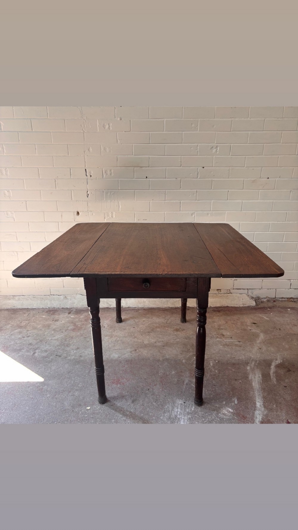 A dark brown wooden drop-leaf table featuring two hinged side leaves, a central drawer with a single knob, and slender turned legs. View II