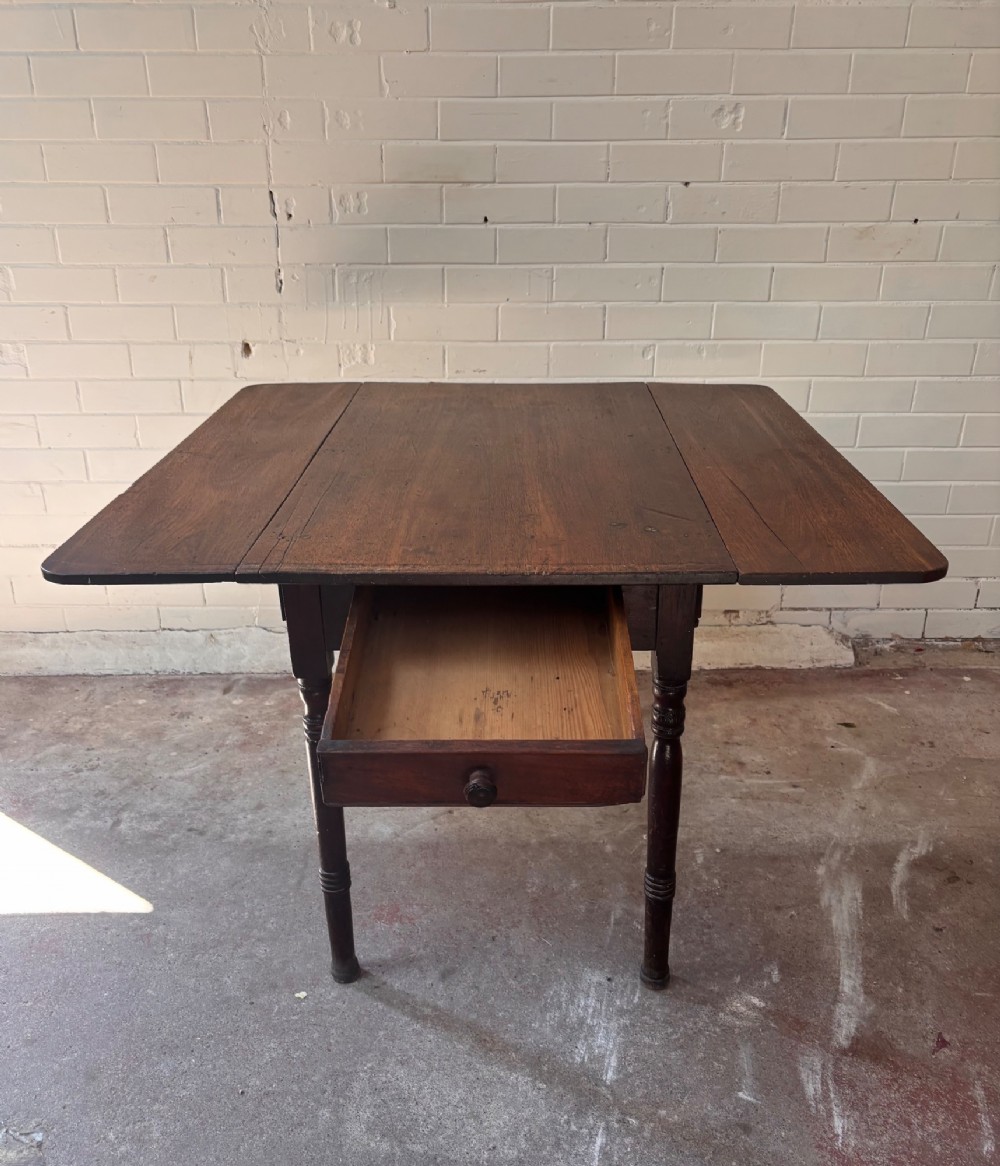 A dark brown wooden drop-leaf table featuring two hinged side leaves, a central drawer with a single knob, and slender turned legs. View III