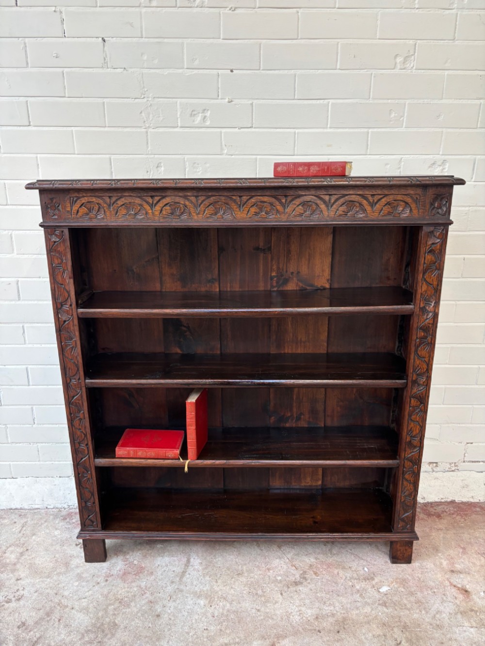 A low dark oak open bookcase featuring detailed lunette carving along the upper frieze and carved foliate borders down the upright sides.
