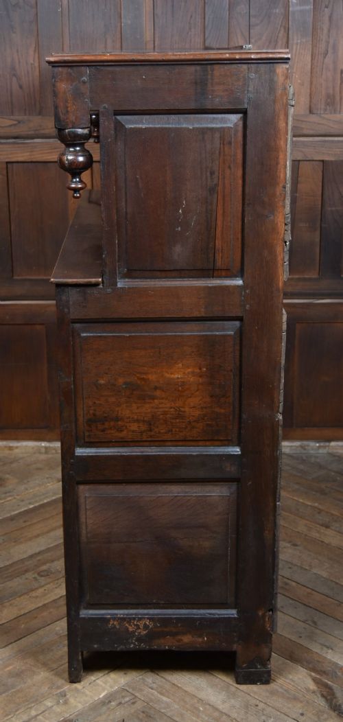 A 17th-century dark oak court cupboard featuring a carved lunette frieze, turned drop pendants, studded panel initials, and geometric lower doors. View VIII
