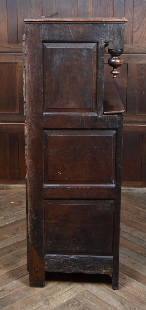 A 17th-century dark oak court cupboard featuring a carved lunette frieze, turned drop pendants, studded panel initials, and geometric lower doors. View X