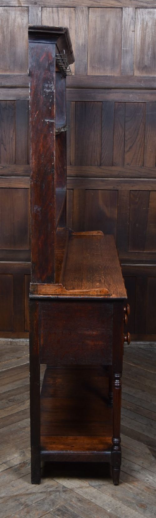 A dark patinated oak country dresser featuring a three-tiered open plate rack stacked with ceramic tableware, set above a base with five drawers, turned front legs, and a low pot board shelf. View XIII