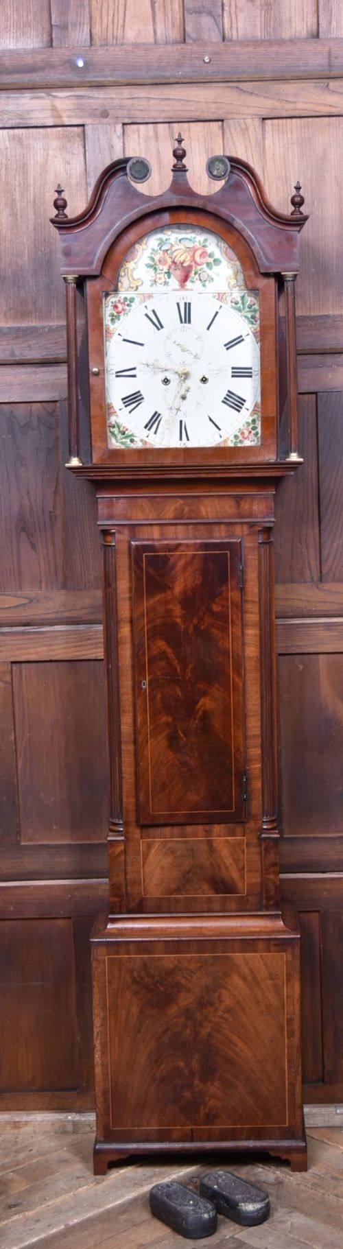 fig 1. A Scottish mahogany longcase clock featuring a swan-neck pediment with brass finials, a hand-painted dial face, and flame mahogany veneers.