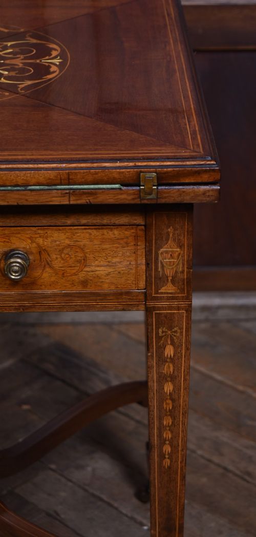 An Edwardian mahogany envelope card table featuring satinwood marquetry inlay, a single drawer, and a curved cross-stretcher with brass castors. View III