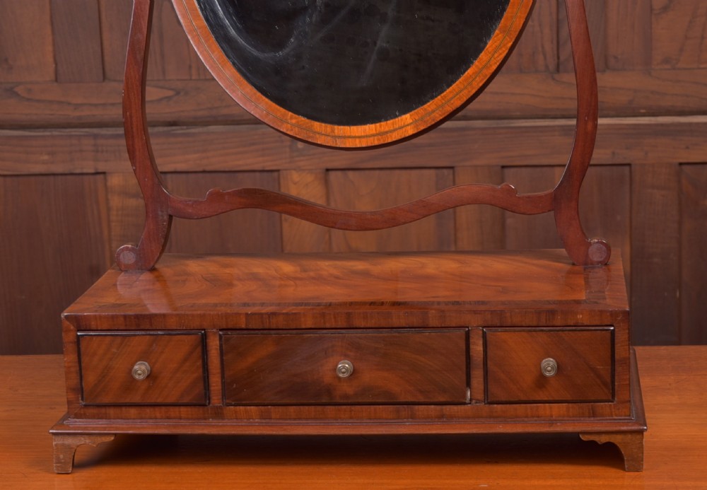 An antique mahogany oval toilet mirror with curved horn supports atop a plinth base containing three small drawers. View II