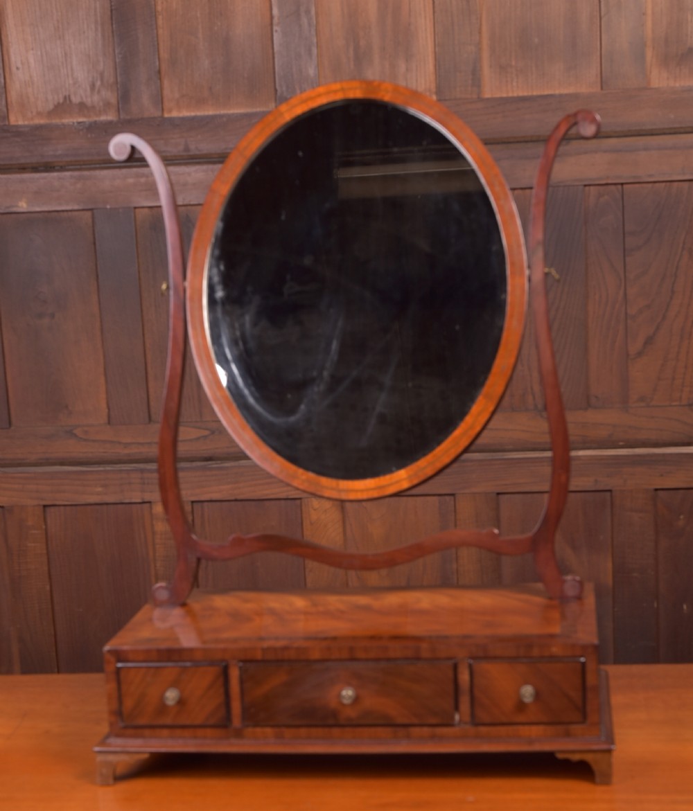 An antique mahogany oval toilet mirror with curved horn supports atop a plinth base containing three small drawers. View IX