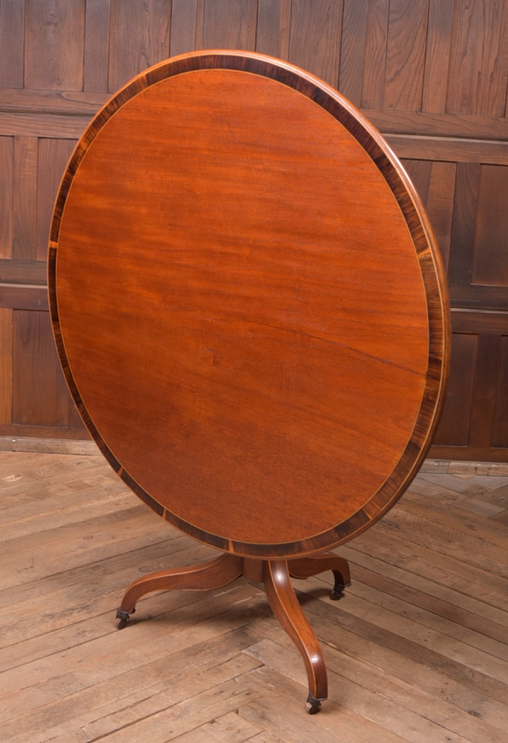 An antique Regency mahogany snap top table with a round crossbanded top, central turned pedestal, four splayed sabre legs, and brass castors.