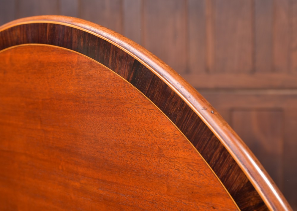 An antique Regency mahogany snap top table with a round crossbanded top, central turned pedestal, four splayed sabre legs, and brass castors. View II