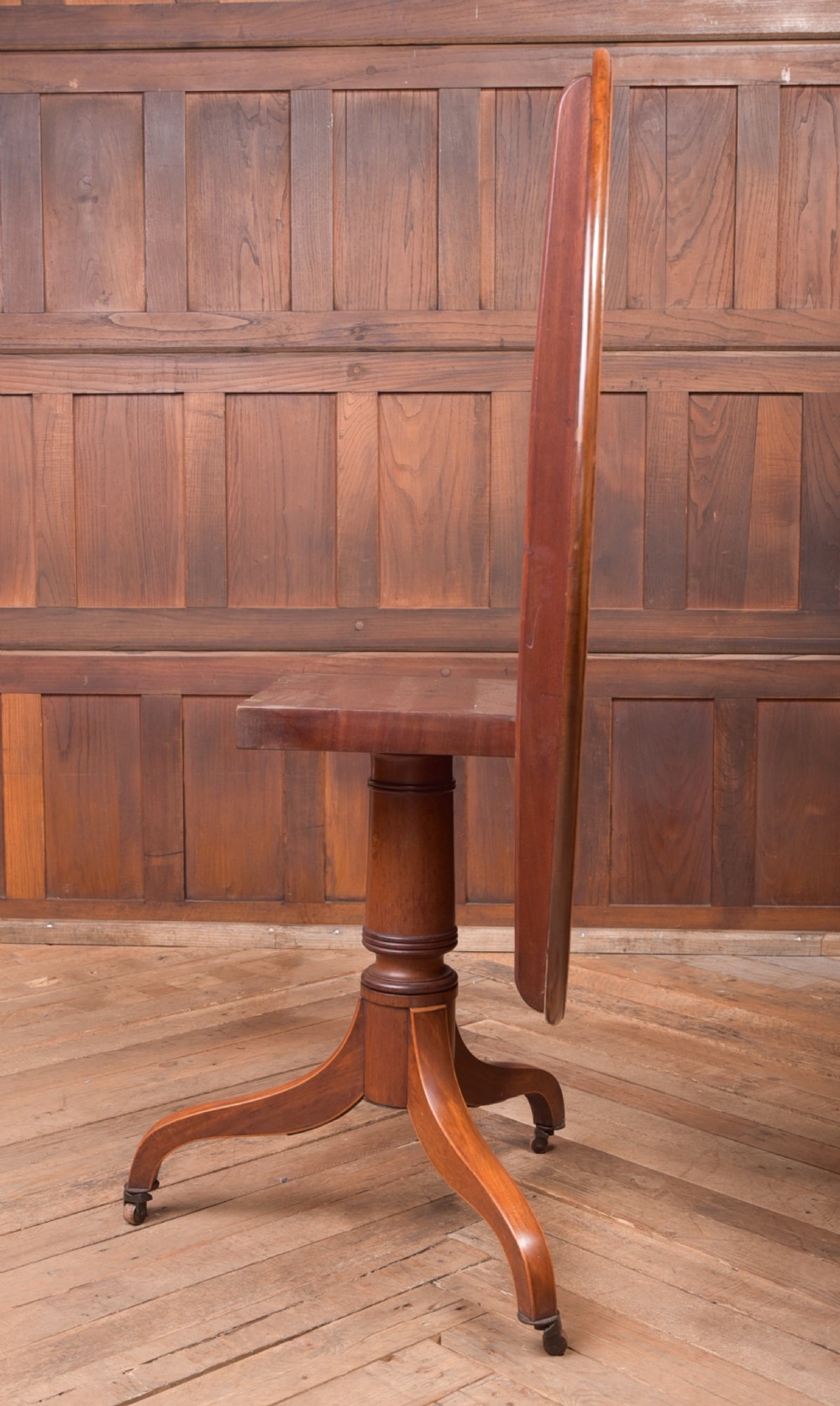 An antique Regency mahogany snap top table with a round crossbanded top, central turned pedestal, four splayed sabre legs, and brass castors. View X
