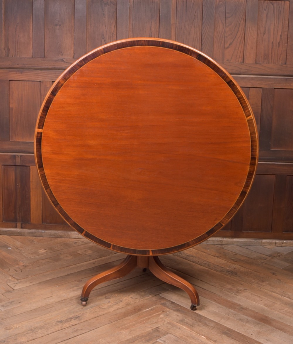 An antique Regency mahogany snap top table with a round crossbanded top, central turned pedestal, four splayed sabre legs, and brass castors. View XI
