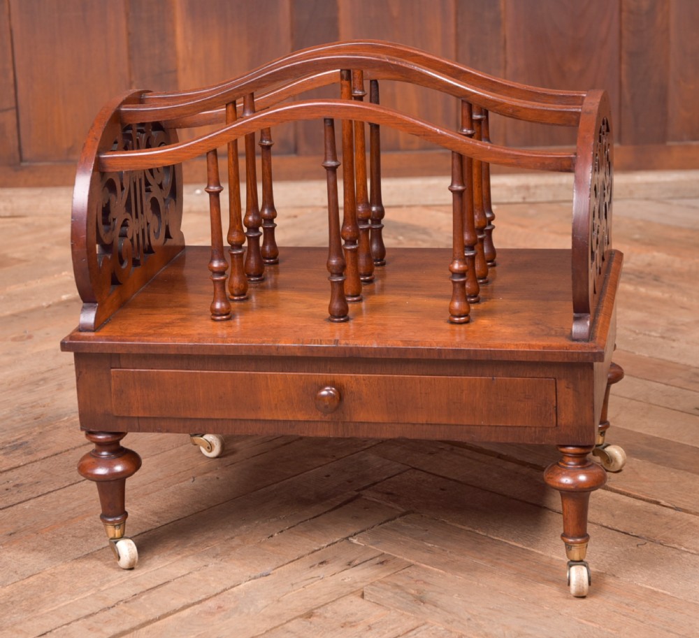 A Victorian walnut Canterbury featuring carved fretwork side panels, turned spindle dividers, a single lower drawer, and turned legs with brass castors.