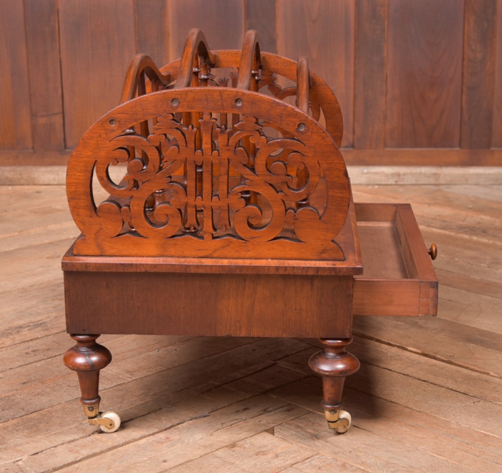 A Victorian walnut Canterbury featuring carved fretwork side panels, turned spindle dividers, a single lower drawer, and turned legs with brass castors. View IV