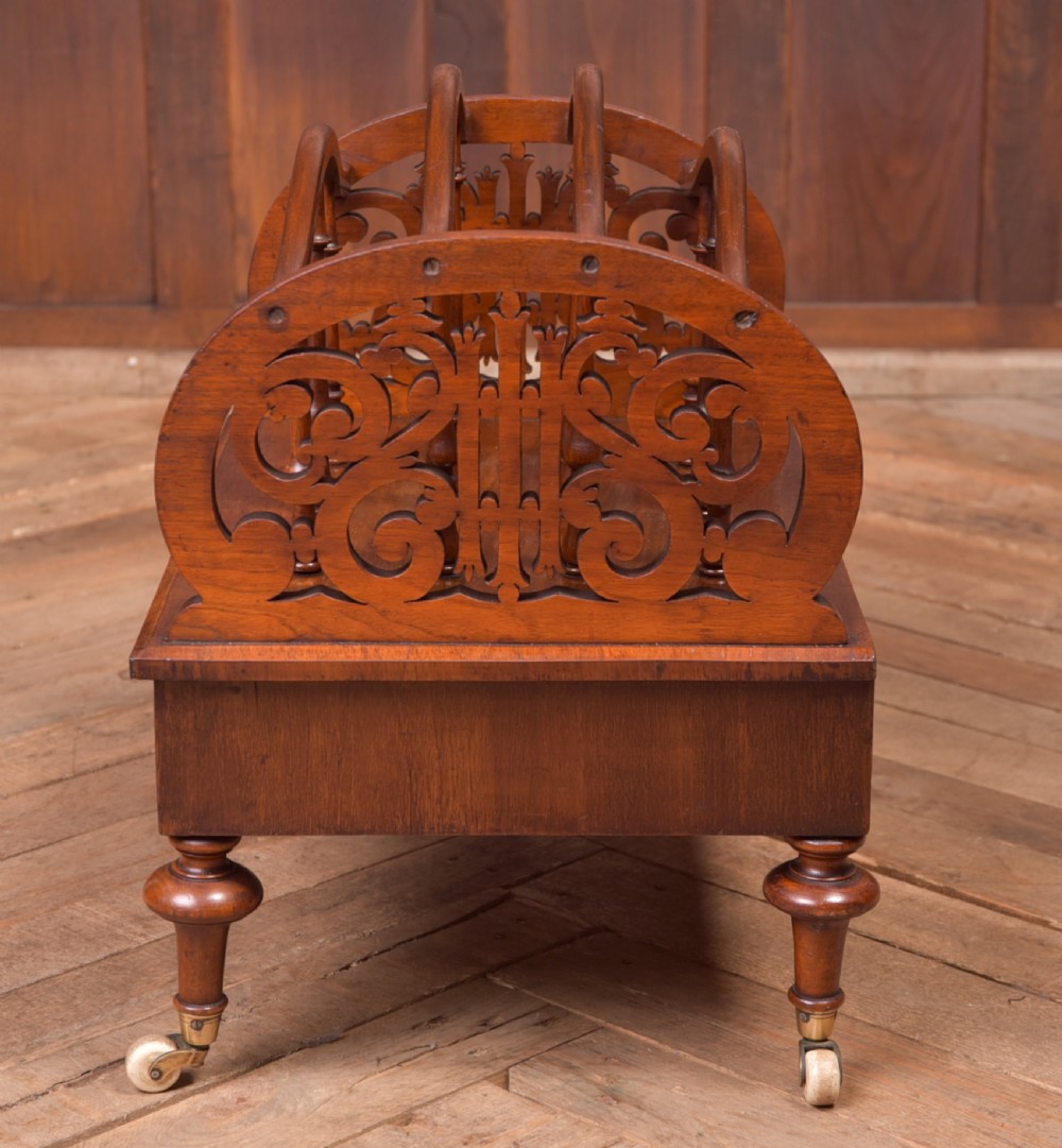 A Victorian walnut Canterbury featuring carved fretwork side panels, turned spindle dividers, a single lower drawer, and turned legs with brass castors. View V