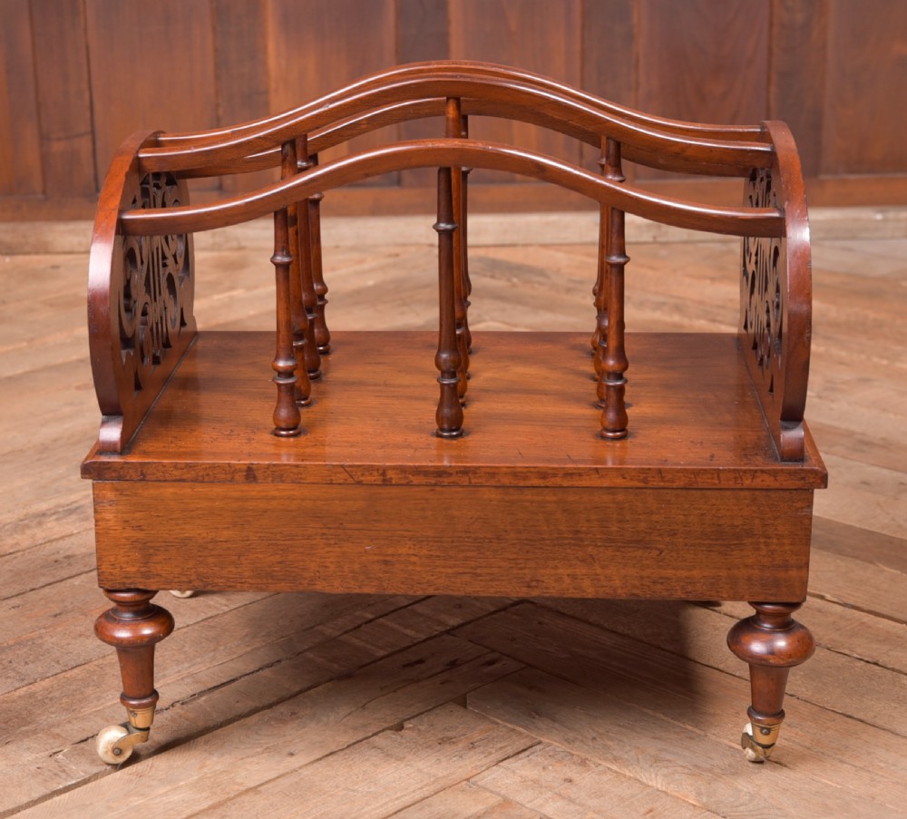 A Victorian walnut Canterbury featuring carved fretwork side panels, turned spindle dividers, a single lower drawer, and turned legs with brass castors. View VI
