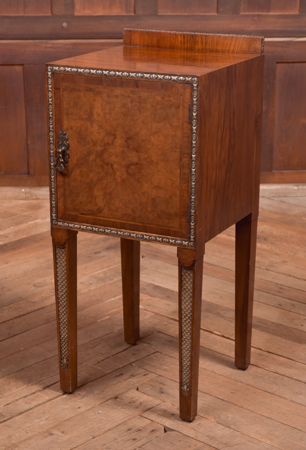 An antique walnut bedside cabinet featuring a single door panel with burr walnut veneer, beaded metal trim, a top splashback upstand, and four square tapered legs.