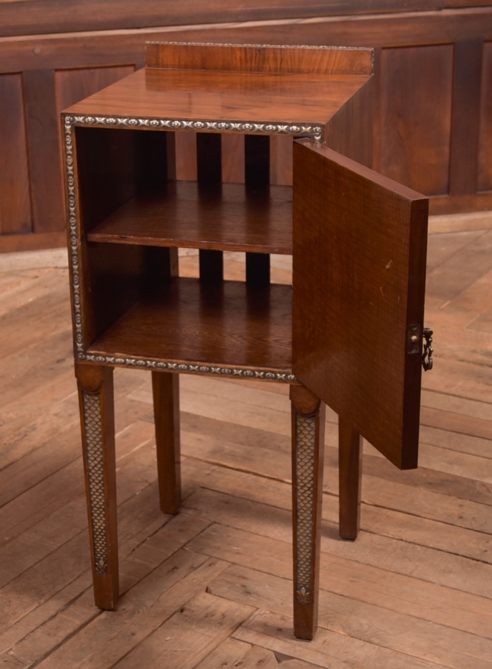 An antique walnut bedside cabinet featuring a single door panel with burr walnut veneer, beaded metal trim, a top splashback upstand, and four square tapered legs. View VI