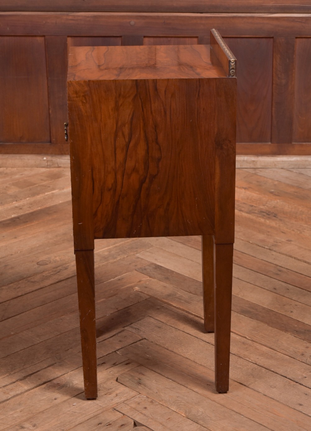 An antique walnut bedside cabinet featuring a single door panel with burr walnut veneer, beaded metal trim, a top splashback upstand, and four square tapered legs. View VII