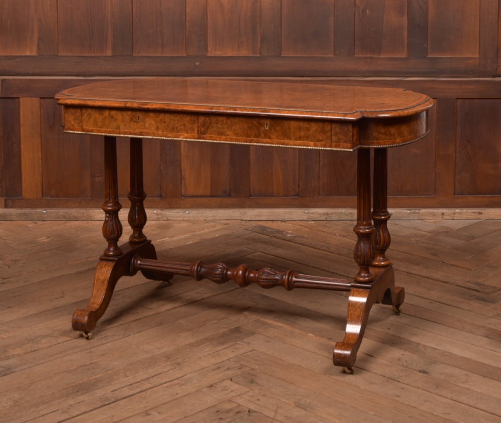 A Victorian burr walnut library table featuring a shaped top, frieze drawers, twin turned legs on a stretchered base, and brass castors.