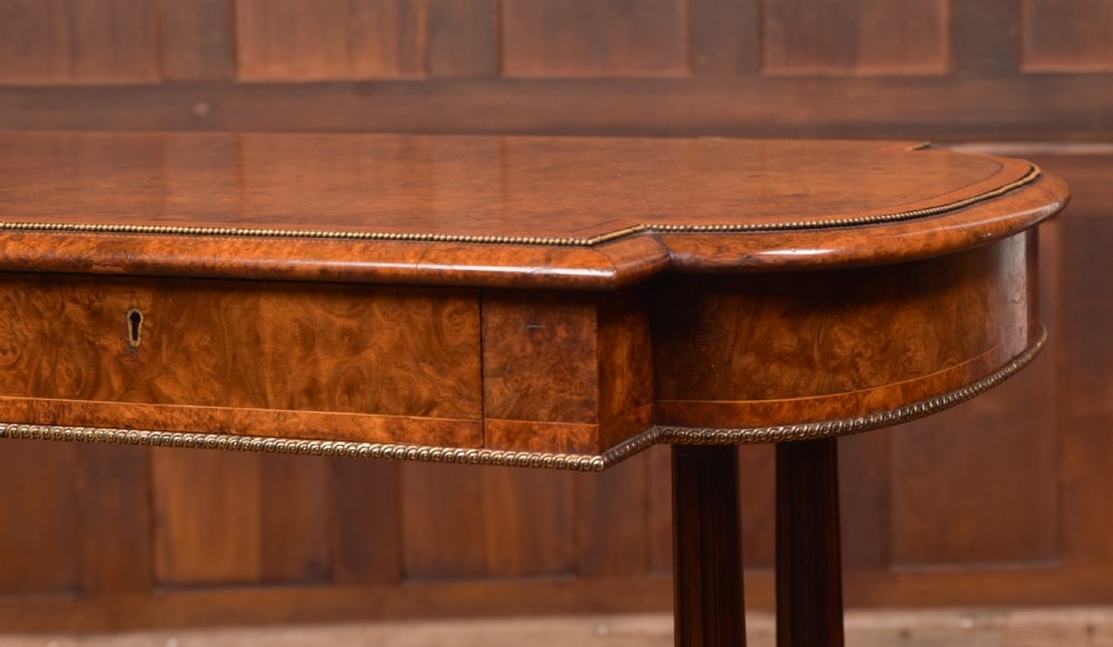A Victorian burr walnut library table featuring a shaped top, frieze drawers, twin turned legs on a stretchered base, and brass castors. View II