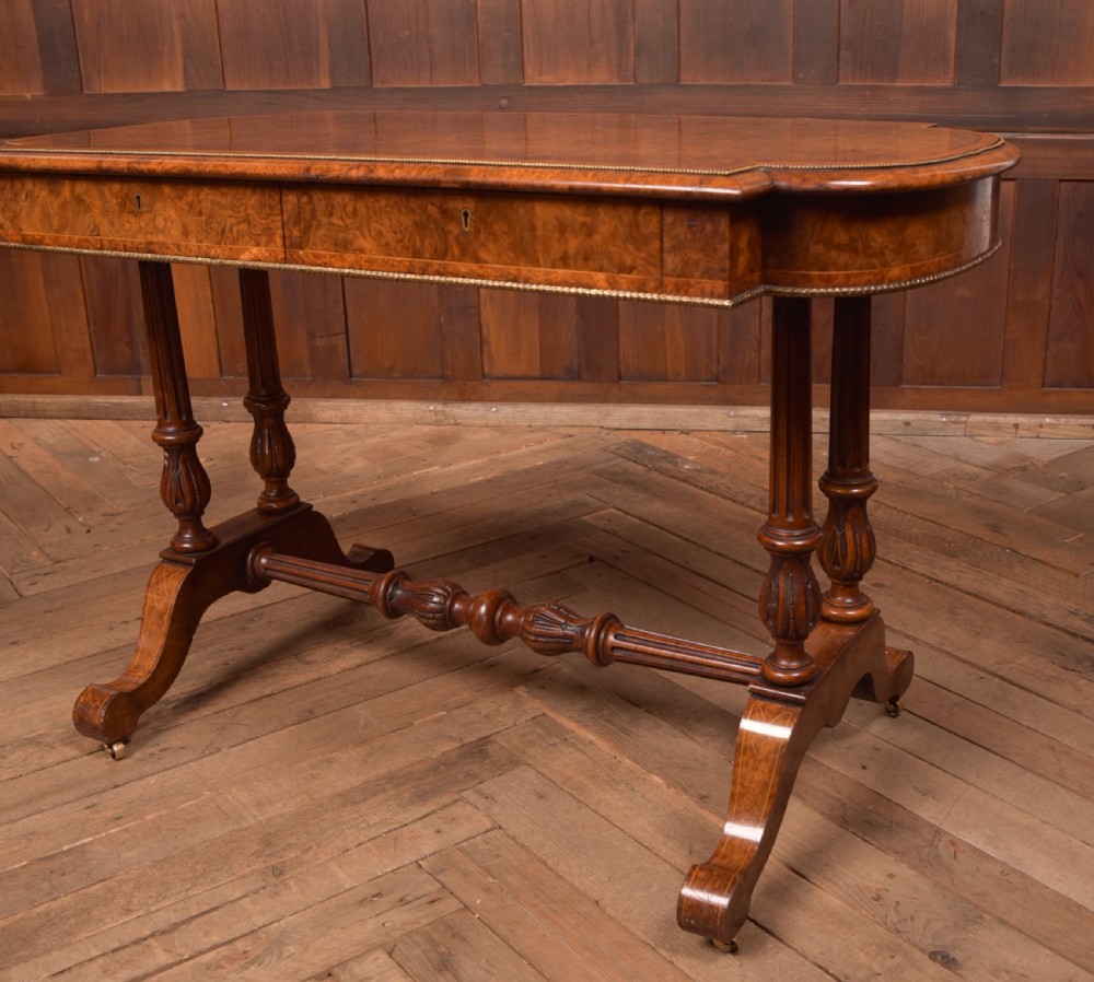 A Victorian burr walnut library table featuring a shaped top, frieze drawers, twin turned legs on a stretchered base, and brass castors. View III