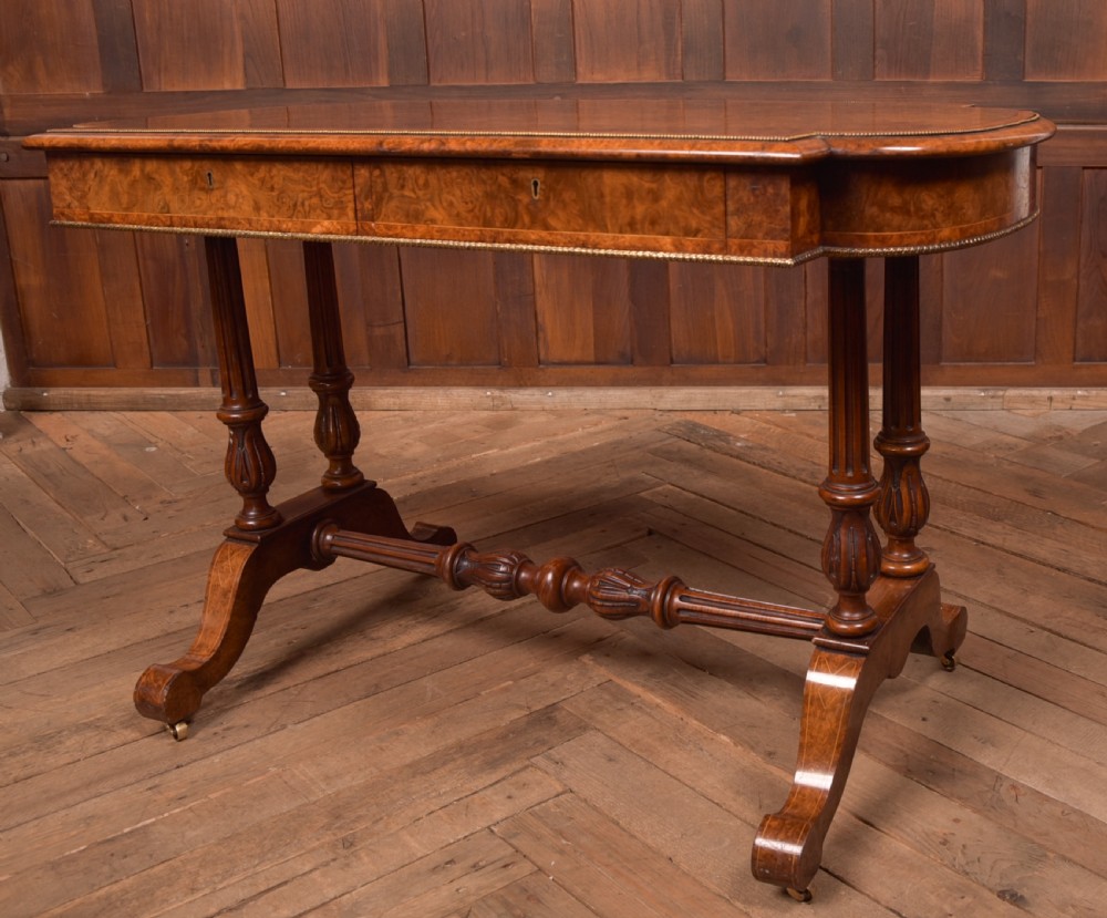 A Victorian burr walnut library table featuring a shaped top, frieze drawers, twin turned legs on a stretchered base, and brass castors. View V