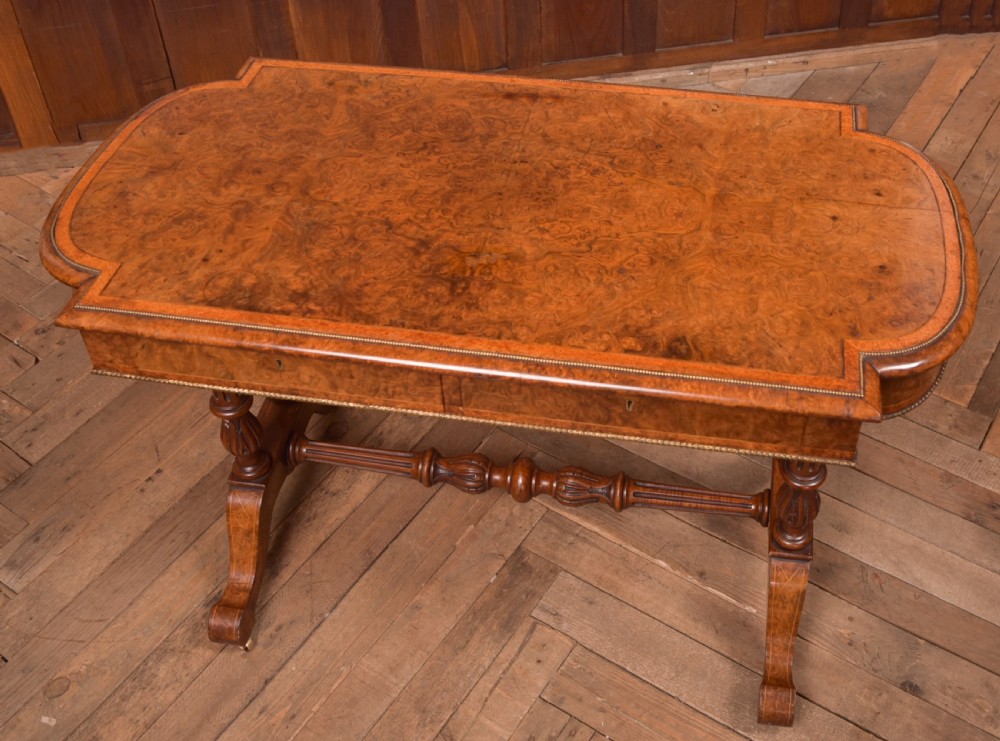 A Victorian burr walnut library table featuring a shaped top, frieze drawers, twin turned legs on a stretchered base, and brass castors. View VI