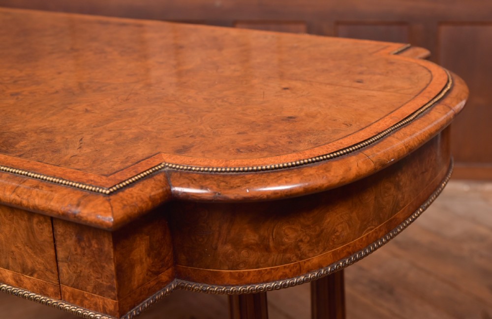 A Victorian burr walnut library table featuring a shaped top, frieze drawers, twin turned legs on a stretchered base, and brass castors. View VII