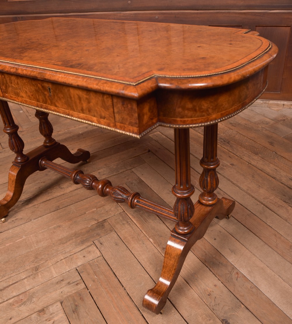 A Victorian burr walnut library table featuring a shaped top, frieze drawers, twin turned legs on a stretchered base, and brass castors. View VIII