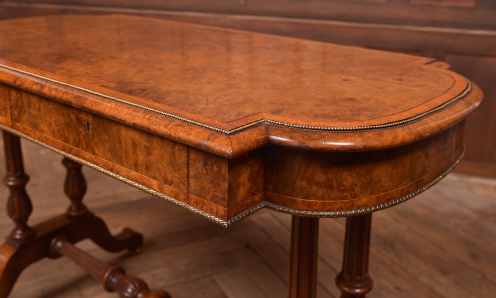 A Victorian burr walnut library table featuring a shaped top, frieze drawers, twin turned legs on a stretchered base, and brass castors. View IX