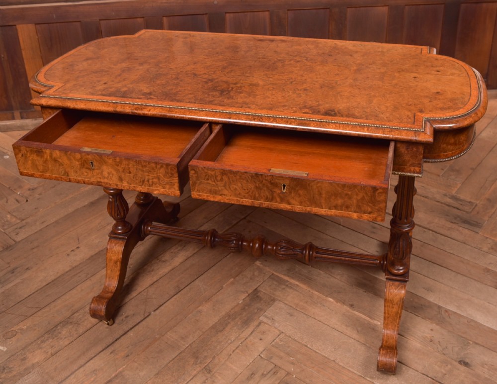 A Victorian burr walnut library table featuring a shaped top, frieze drawers, twin turned legs on a stretchered base, and brass castors. View X