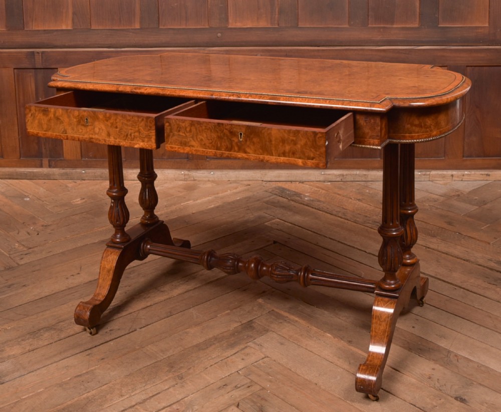 A Victorian burr walnut library table featuring a shaped top, frieze drawers, twin turned legs on a stretchered base, and brass castors. View XI