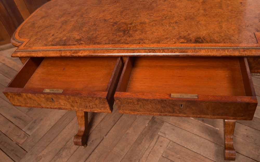 A Victorian burr walnut library table featuring a shaped top, frieze drawers, twin turned legs on a stretchered base, and brass castors. View XII