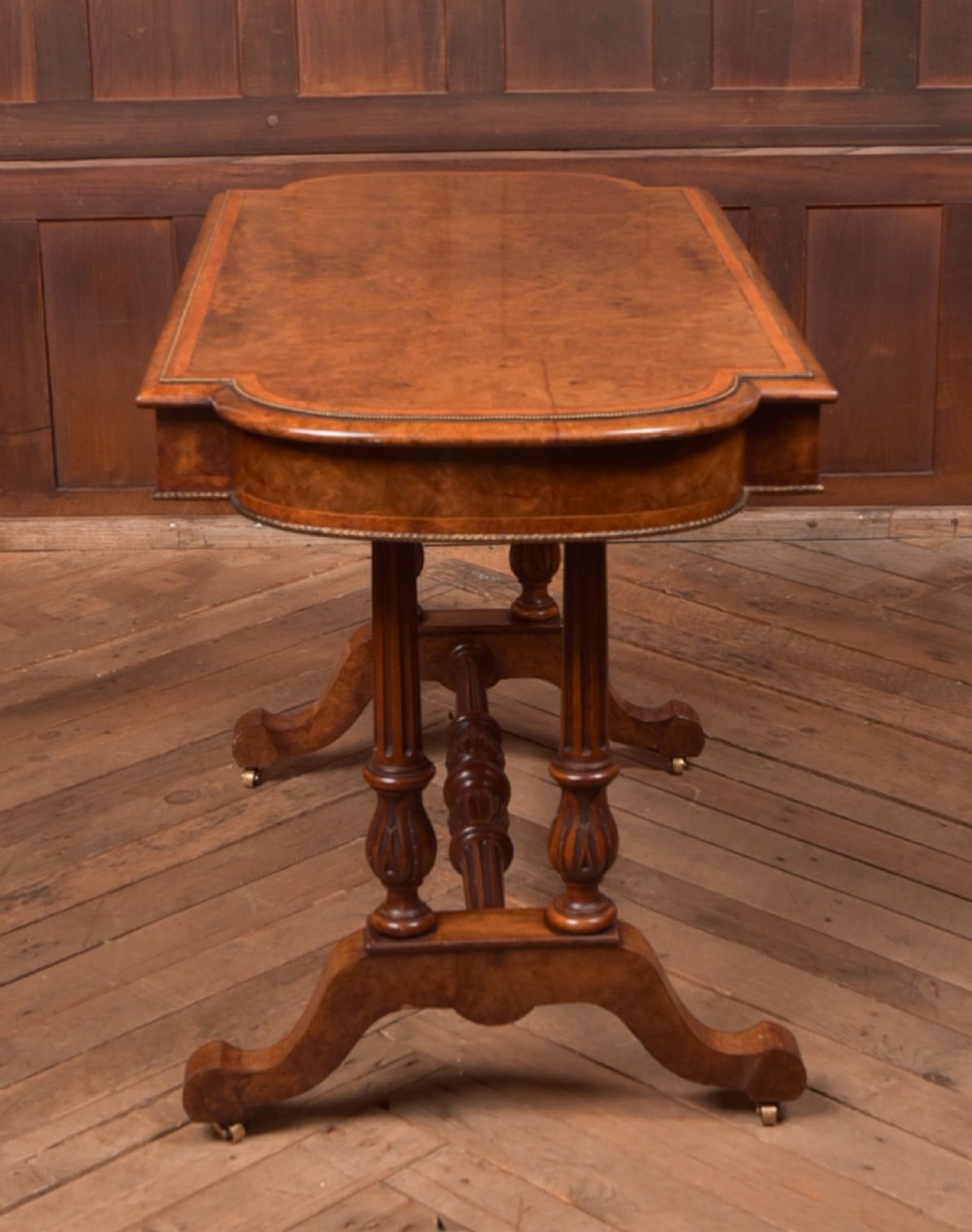 A Victorian burr walnut library table featuring a shaped top, frieze drawers, twin turned legs on a stretchered base, and brass castors. View XIII