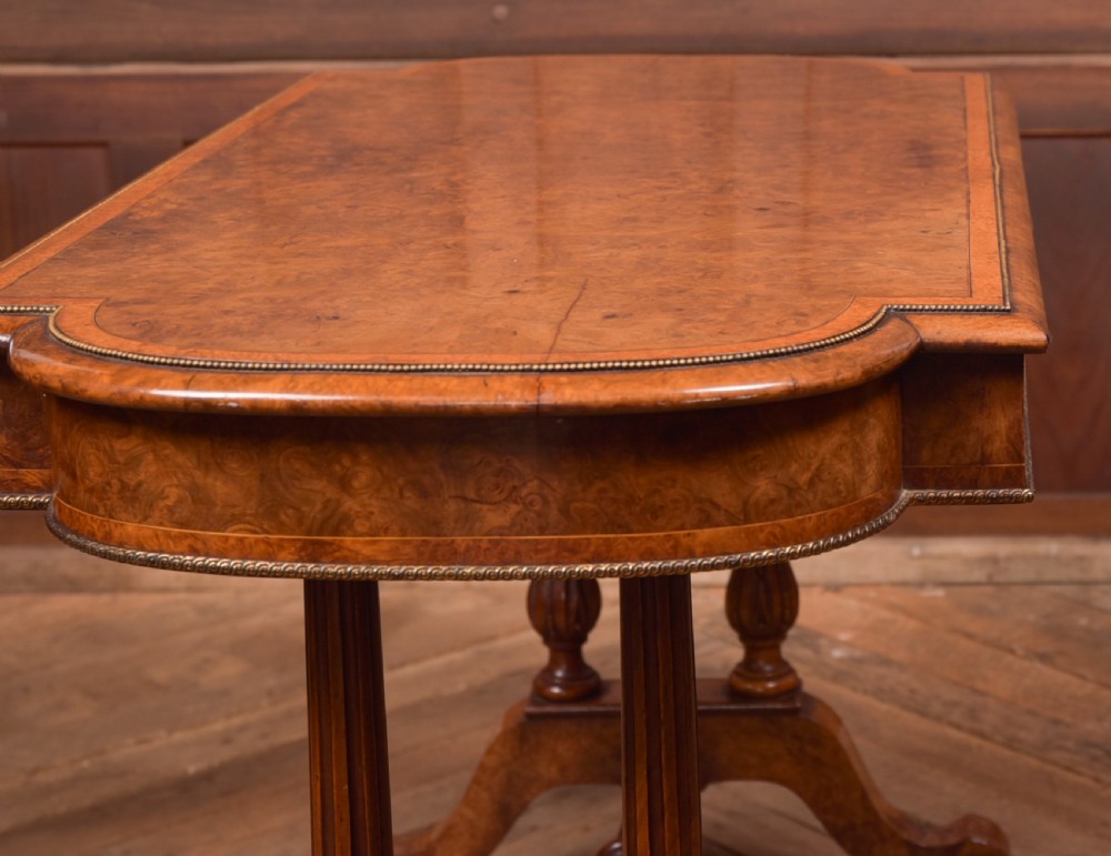 A Victorian burr walnut library table featuring a shaped top, frieze drawers, twin turned legs on a stretchered base, and brass castors. View XIV