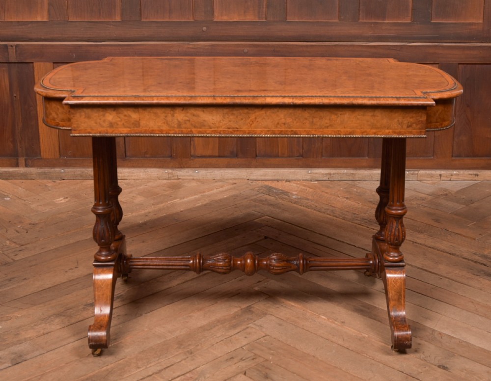 A Victorian burr walnut library table featuring a shaped top, frieze drawers, twin turned legs on a stretchered base, and brass castors. View XV