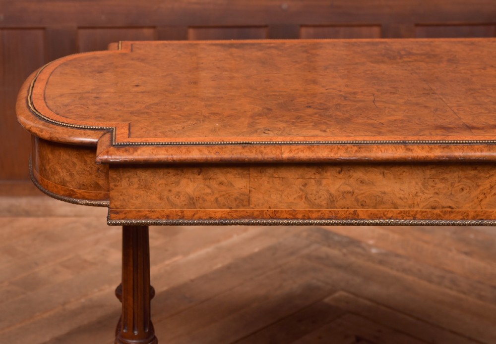 A Victorian burr walnut library table featuring a shaped top, frieze drawers, twin turned legs on a stretchered base, and brass castors. View XVI