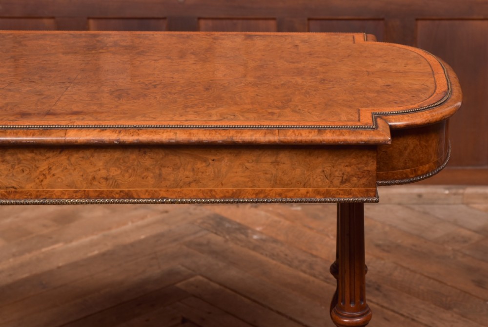 A Victorian burr walnut library table featuring a shaped top, frieze drawers, twin turned legs on a stretchered base, and brass castors. View XVII