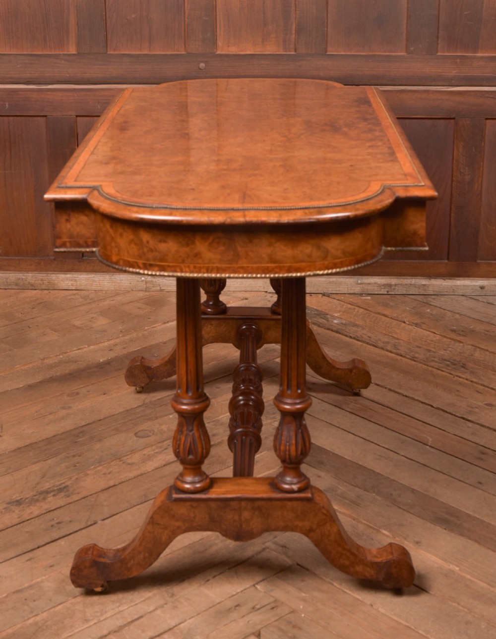A Victorian burr walnut library table featuring a shaped top, frieze drawers, twin turned legs on a stretchered base, and brass castors. View XVIII