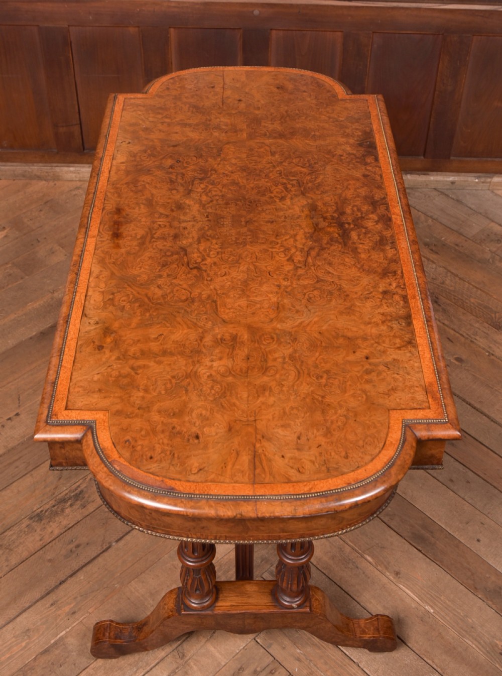 A Victorian burr walnut library table featuring a shaped top, frieze drawers, twin turned legs on a stretchered base, and brass castors. View XIX