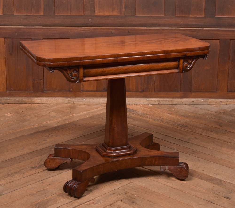 A Victorian mahogany card table featuring a rectangular fold-over top set upon a central tapered pedestal and a quadripartite plinth base with carved feet.