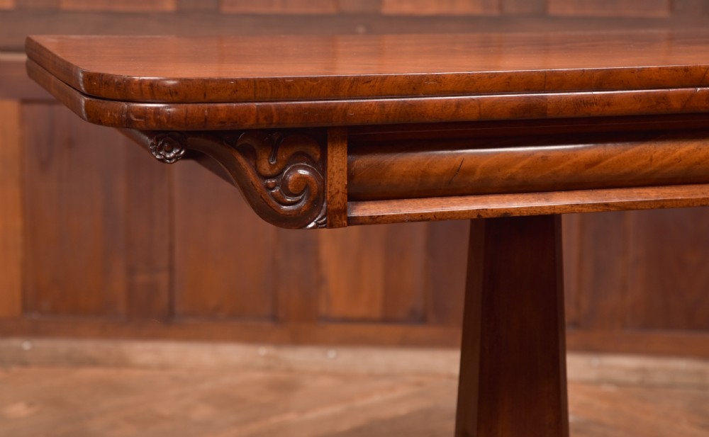 A Victorian mahogany card table featuring a rectangular fold-over top set upon a central tapered pedestal and a quadripartite plinth base with carved feet. View II