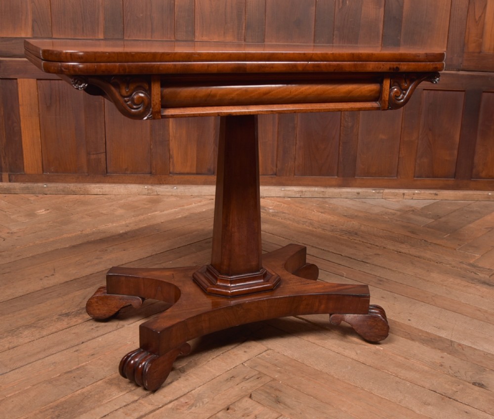 A Victorian mahogany card table featuring a rectangular fold-over top set upon a central tapered pedestal and a quadripartite plinth base with carved feet. View V