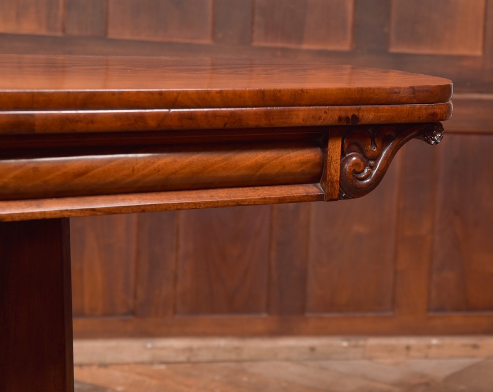 A Victorian mahogany card table featuring a rectangular fold-over top set upon a central tapered pedestal and a quadripartite plinth base with carved feet. View VI