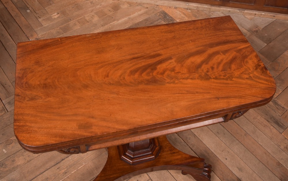 A Victorian mahogany card table featuring a rectangular fold-over top set upon a central tapered pedestal and a quadripartite plinth base with carved feet. View VIII