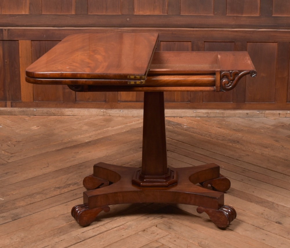 A Victorian mahogany card table featuring a rectangular fold-over top set upon a central tapered pedestal and a quadripartite plinth base with carved feet. View IX