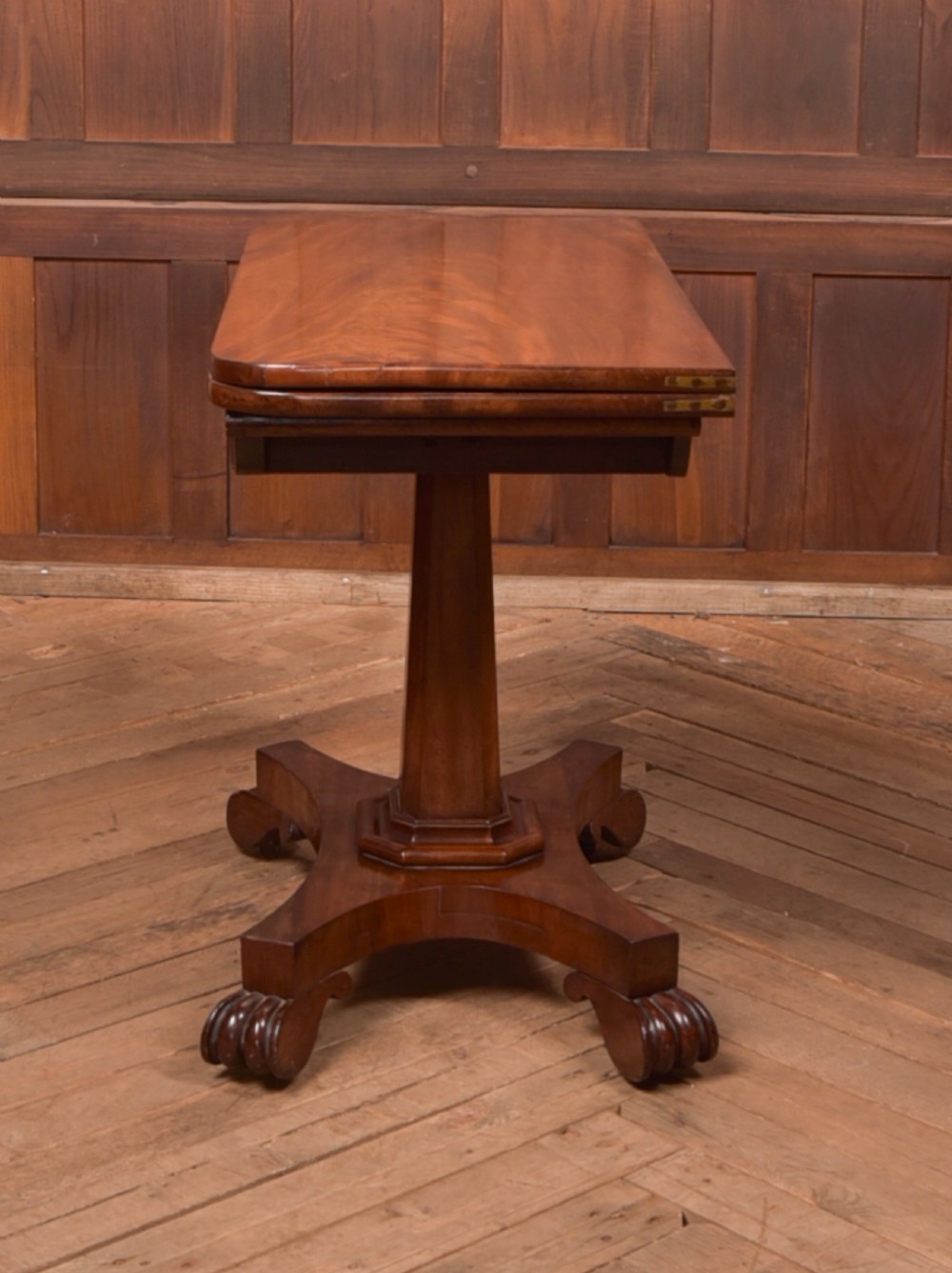 A Victorian mahogany card table featuring a rectangular fold-over top set upon a central tapered pedestal and a quadripartite plinth base with carved feet. View XIII