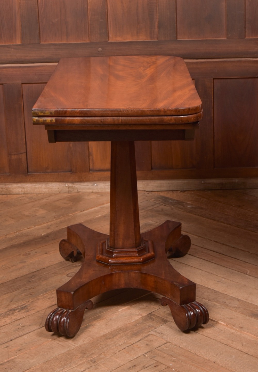A Victorian mahogany card table featuring a rectangular fold-over top set upon a central tapered pedestal and a quadripartite plinth base with carved feet. View XV